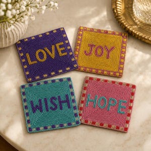 May include: Four square coasters crafted from small beads. The purple coaster features the word LOVE in gold. The yellow coaster displays the word JOY in pink. The teal coaster shows the word WISH in purple. The pink coaster features the word HOPE in teal.