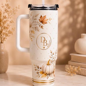 Peut inclure: Un gobelet isotherme couleur crème avec une poignée, un couvercle transparent et une paille. L'extérieur présente un motif en losanges doré, des feuilles d'automne, une citrouille blanche et un monogramme doré avec les lettres DB et un cœur.