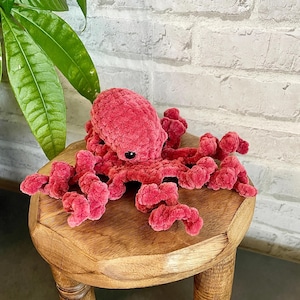 Könnte beinhalten: Ein Plüsch-Oktopus aus weichem, strukturiertem dunkelrosa Garn. Der Oktopus hat einen abgerundeten Kopf, ein einzelnes sichtbares schwarzes Kunststoffauge und acht gekräuselte, tentakelartige Arme, die sich über die Oberfläche erstrecken.