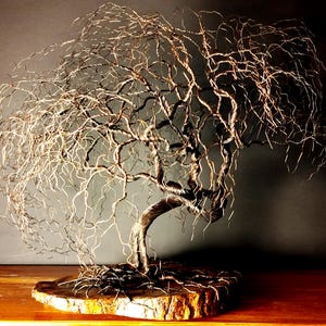 May include: A sculpture of a weeping willow tree crafted from thin, dark metallic wire. The tree features a twisted trunk and numerous delicate, drooping branches that spread outward. It is mounted on a circular, natural-edged wooden base.