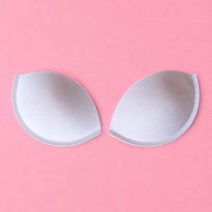 May include: A pair of smooth, light grey, semi-circular foam bra inserts. These moulded cups feature a rounded, teardrop-like shape with a clean, finished edge, designed to provide structure and padding for garments, presented as two separate pieces.