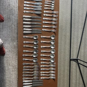 Peut inclure: Une grande collection de couverts argentés disposés en rangées, comprenant des couteaux, des fourchettes et des cuillères de différentes tailles. L'ensemble présente des manches décoratifs ornés avec une finition métallique uniforme.