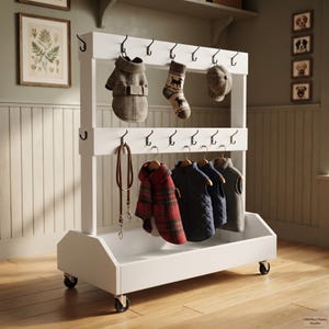 Puede incluir: Un perchero de almacenamiento blanco con ruedas y dos barras horizontales con múltiples ganchos de metal negro. El estante sostiene un abrigo beige para perro, un calcetín estampado, una gorra, una correa de cuero marrón, un abrigo de cuadros rojos y dos chalecos acolchados en perchas.