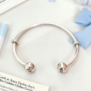 May include: A silver-toned open bangle bracelet featuring two spherical end caps. Each sphere is engraved with black lettering, with one side displaying the letters A, O, and P, and the other side showing the letters D, O, and R.