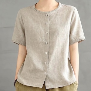 May include: A short-sleeved beige blouse featuring a round neckline with a subtle ruffled trim. The garment has a front button-down closure with seven light-colored buttons running vertically down the center. The sleeves also feature a ruffled edge.