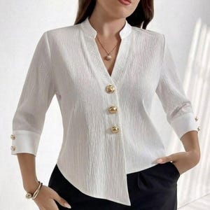 Puede incluir: Una blusa blanca texturizada con cuello en V y solapa lisa, mangas tres cuartos con puños blancos y una tapeta frontal adornada con tres botones grandes, redondos y dorados. La tela presenta una sutil textura vertical arrugada.