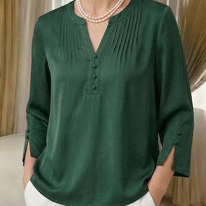 May include: A dark green long-sleeve blouse featuring a V-neckline with a vertical row of four matching fabric-covered buttons. The garment includes delicate pleating details along the shoulders and chest, with two small buttons on each sleeve cuff.