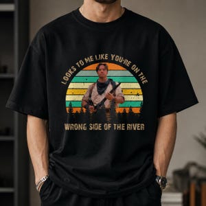 May include: A black t-shirt featuring a graphic print of a person holding a rifle, set against a sunset background with silhouettes of pine trees. The text reads, "LOOKS TO ME LIKE YOU'RE ON THE WRONG SIDE OF THE RIVER" in a distressed font.