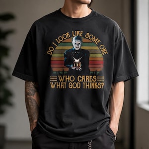 May include: A black short-sleeve t-shirt featuring a graphic of Pinhead holding a puzzle box against a retro sunset background. The text reads: DO I LOOK LIKE SOME ONE WHO CARES WHAT GOD THINKS? The design uses yellow, orange, and red tones.