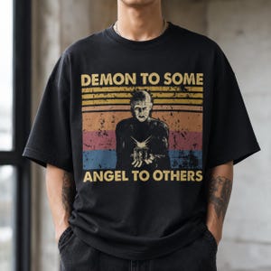 May include: A black short-sleeve t-shirt featuring a graphic print of a figure holding a puzzle box. The design includes horizontal stripes in yellow, orange, pink, and blue, with the text "DEMON TO SOME" above and "ANGEL TO OTHERS" below.