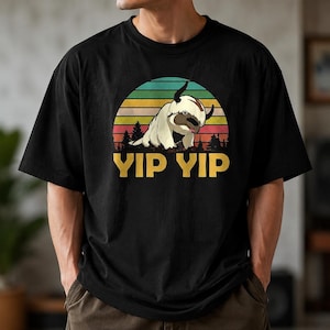 May include: A black t-shirt featuring a graphic of a cream-colored, six-legged creature with horns. The design includes a sunset background with horizontal stripes in green, yellow, orange, and red, and the text YIP YIP printed in yellow below.