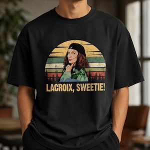 May include: A black crew neck t-shirt featuring a graphic print of a person wearing a black beret. The design includes a sunset background with horizontal stripes and the text LACROIX, SWEETIE! printed in yellow capital letters below the image.