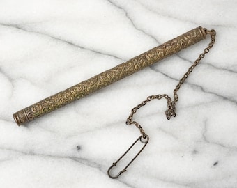 May include: A long, cylindrical brass needle case featuring an intricate, embossed floral pattern across its entire surface. A thin, matching metal chain is attached to one end of the tube, connecting to a simple metal safety pin at the other end.