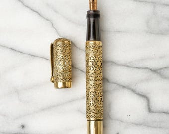 May include: A fountain pen with a gold-colored metal barrel and cap featuring an intricate filigree pattern. The pen has a black grip section, a gold-colored nib, and a matching metal clip on the cap. The design consists of repeating geometric shapes.