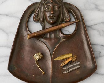 May include: A bronze-toned metal desk tray featuring a sculpted female face with a floral headband. The tray holds a long, slender metal pen, a small ornate metal ring with a dangling ball, and four assorted metal calligraphy nibs of varying colors.