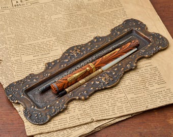 May include: An ornate, rectangular bronze-toned metal pen tray with embossed floral and scrollwork details. Resting inside the tray are two writing instruments: one fountain pen with a marbled orange and black pattern and a gold clip, and a slender pen.