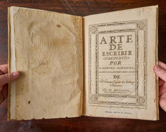 May include: An open antique book with aged, cream-colored paper pages. The right page features a decorative border and black printed text that reads: ARTE DE ESCRIBIR COMPUESTO POR D. ESTEVAN XIMENEZ, AÑO DE MDCCLXXXIX, along with additional publication details.