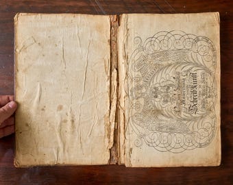 May include: An antique open book with weathered, cream-colored pages and a damaged spine. The right page features intricate black ink calligraphy and a detailed illustration of winged figures surrounded by ornate, swirling decorative line work.