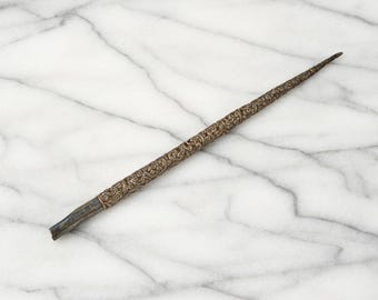 May include: A long, slender, tapered rod featuring an intricate, embossed floral scrollwork pattern across its entire length. The object has a metallic, bronze-toned finish with a dark, smooth, unadorned section at the base end, tapering to a point.