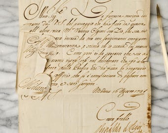 May include: An antique, off-white parchment document featuring ornate, dark brown cursive calligraphy in Italian. The paper shows visible creases, frayed edges, and a small tear on the left side. A thin, light-colored quill pen rests to the right.