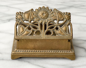 May include: A rectangular brass desk accessory featuring an openwork top with a central sunflower motif and stylized leaves. The base has a beaded border detail along the bottom edge, and the entire piece has a muted, aged metallic finish.