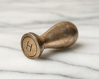 May include: A brass wax seal stamp featuring a rounded handle and a circular flat base. The face of the stamp is engraved with the capital letter H centered within a circular border. The metal surface shows a weathered, aged patina throughout.