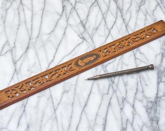 May include: A long, rectangular wooden ruler featuring intricate floral cutouts and a central oval inlay depicting a figure. Beside the ruler lies a slim, textured metallic mechanical pencil with a dark finish and a small clip at the top end.