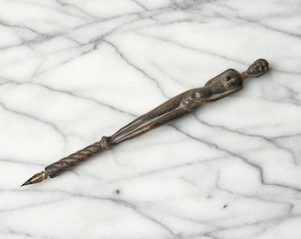 May include: A dark brown carved wooden dip pen featuring a slender human figure forming the handle, with a twisted rope-like design extending down to a pointed metal nib at the tip. The pen has a smooth, polished finish and elongated proportions.