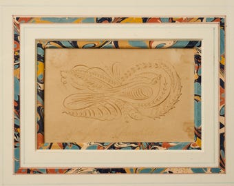 May include: A framed calligraphic drawing on light tan paper featuring a stylized bird surrounded by intricate swirling flourishes and leaf patterns. The frame has a decorative border with a marbled pattern in shades of blue, orange, and yellow.