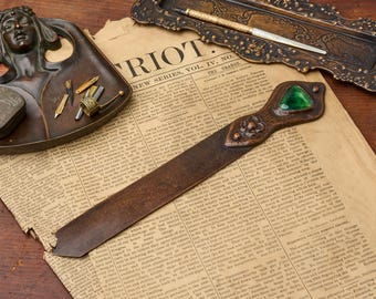 May include: A metal letter opener with a dark, weathered finish featuring a triangular green gemstone at the handle and a small embossed floral detail. The blade is flat and elongated, tapering slightly toward the pointed tip, showcasing a vintage aesthetic.