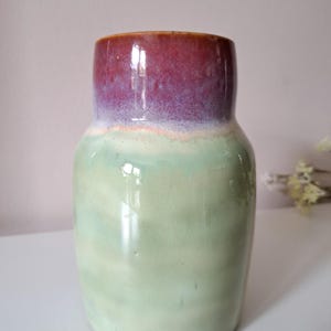 Può includere: Un vaso in ceramica di forma cilindrica con una finitura liscia e lucida. La parte superiore presenta uno smalto color magenta intenso e viola, mentre il corpo inferiore è di un tenue verde menta chiaro. I colori sono sfumati tra loro.