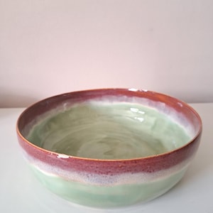 Puede incluir: Un cuenco de cerámica redondo con un interior liso de color verde claro y un borde ancho de color marrón rojizo oscuro. El exterior muestra una transición degradada desde el borde superior marrón rojizo oscuro hasta una base verde pálido.