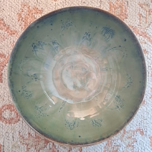 Può includere: Una ciotola rotonda in ceramica con una smaltatura liscia e lucida nei toniida nei toni del verde salvia e del beige tenue. La superficie interna presenta un motivo radiale di pennellate maculate blu scuro che si irradiano dal centro verso il bordo.