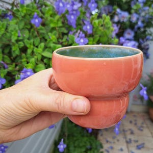 Op de afbeelding: Een kleine, ronde keramische beker met een gladde, glanzende koraalroze buitenkant en een contrasterende diepblauwe glazuurlaag aan de binnenkant. De beker heeft een duidelijke horizontale inkeping rond het midden van de vorm.
