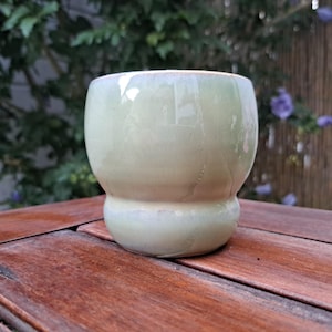 Op de afbeelding: Een kleine, ronde keramische beker met een glanzend, bleek saliegroen glazuur. De beker heeft een duidelijk breder bovendeel dat taps toeloopt naar een smallere, bolvormige basis, wat zorgt voor een glad en doorlopend silhouet.