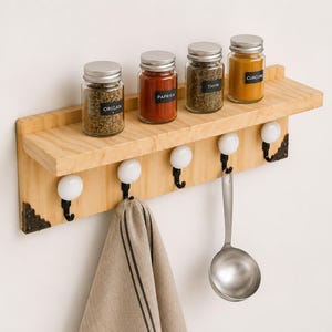 May include: A light-colored wooden wall shelf featuring five black metal hooks with white ceramic knobs. The shelf holds four glass spice jars labeled Origan, Paprika, Thym, and Curcuma. A beige kitchen towel and a metal ladle hang from the hooks.