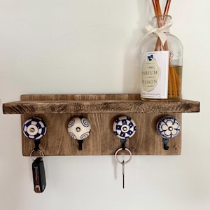 May include: A rectangular wooden wall shelf with a dark brown finish featuring four black metal hooks topped with round ceramic knobs. Each knob displays a unique blue and white pattern. A glass bottle with a white label reading Parfum Maison sits on top.