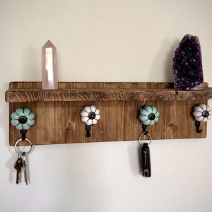 Peut inclure: Une étagère murale en bois dotée de quatre boutons décoratifs en forme de citrouille bleu clair et blanc, chacun avec un crochet en métal noir en dessous. Une pointe de cristal rose clair et un amas d'améthyste violet foncé sont posés dessus.