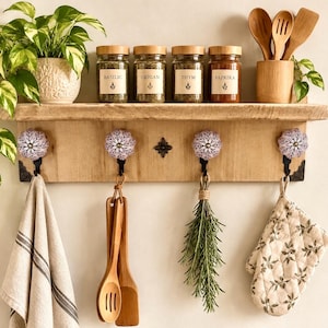 Può includere: Una mensola da parete in legno chiaro con quattro ganci in metallo nero dotati di pomelli floreali color lavanda. La mensola ospita quattro barattoli di spezie in vetro etichettati Basilic, Origan, Thym e Paprika, un portautensili in bambù e una pianta.