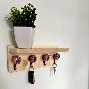 Peut inclure: Une étagère murale en bois clair dotée de quatre crochets en métal noir, chacun surmonté d'un bouton en céramique mauve. Un pot carré blanc contenant une plante artificielle verte est posé sur l'étagère, et deux trousseaux de clés sont suspendus.