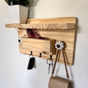 Puede incluir: Un organizador de pared de madera clara con un estante superior, un compartimento de almacenamiento central y tres ganchos de metal negro. La unidad está decorada con dos pomos de cerámica blanca en forma de calabaza.