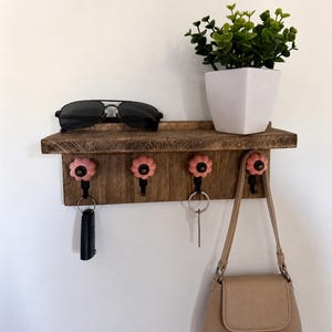 Peut inclure: Une étagère murale en bois brun foncé dotée de quatre crochets en métal noir surmontés de boutons en céramique rose en forme de fleur. L'étagère porte une paire de lunettes de soleil noires et un pot carré blanc avec une plante verte, des clés et un sac beige.