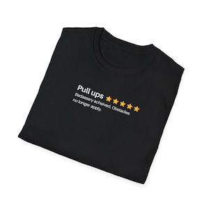 May include: A black t-shirt featuring white text that reads Pull ups followed by five yellow stars and the text Badassery acheived. Obstacles no longer apply. The shirt is folded and displayed against a plain white background for clear visibility.