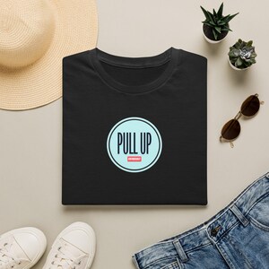 Peut inclure: Un t-shirt noir à manches courtes avec un motif circulaire bleu clair sur la poitrine. À l'intérieur du cercle, le texte PULL UP est écrit en lettres capitales noires, avec le mot ENTHUSIAST en blanc sur un petit rectangle rouge en dessous.