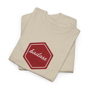 Può includere: Una t-shirt beige a maniche corte con una grafica esagonale rossa sul petto. All'interno dell'esagono, la parola "badass" è scritta in corsivo bianco. La maglietta è piegata e mostra un girocollo e una consistenza del tessuto liscia.