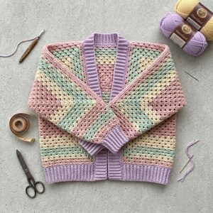 May include: A handmade crochet cardigan featuring a granny square pattern in pastel shades of lavender, mint green, and pale yellow. The garment has long sleeves, a ribbed lavender trim along the front opening, and matching ribbed cuffs at the wrists.