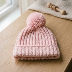 May include: A light pink knit beanie featuring a ribbed texture and a matching pom-pom on top. The hat is constructed with a thick, folded brim and a consistent vertical stitch pattern throughout the crown, providing a soft and uniform appearance.