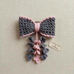 May include: A crocheted bow in dark gray with light pink trim and a central knot. Three dangling strands of small crocheted leaves in alternating gray and pink hang from the center. A small rectangular tag reads Crochet Bow Pattern by Crochevialoop.