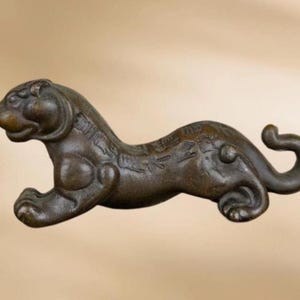 May include: A bronze-toned metal figurine of a crouching tiger in a low, elongated posture. The surface features a dark, weathered patina with visible engraved Chinese characters along the back and side, and a curved tail extending upwards.