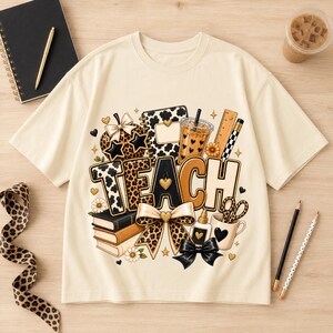 May include: A cream-colored t-shirt featuring a graphic print with the word TEACH in large letters. The design includes illustrations of books, a leopard print bow, a coffee cup, a ruler, a pencil, scissors, and small gold heart accents.